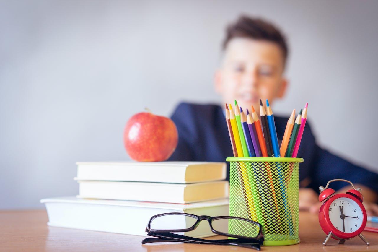 a child sitting at a desk with books, pencils, glasses, and an apple in the foreground