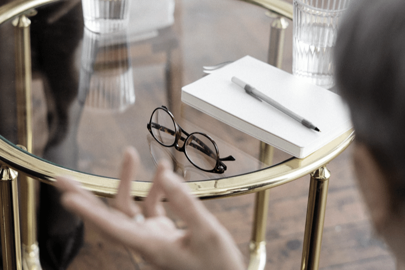 a person holding her hand out in front of a pair of glasses and a blank notepad and pen on a glass table