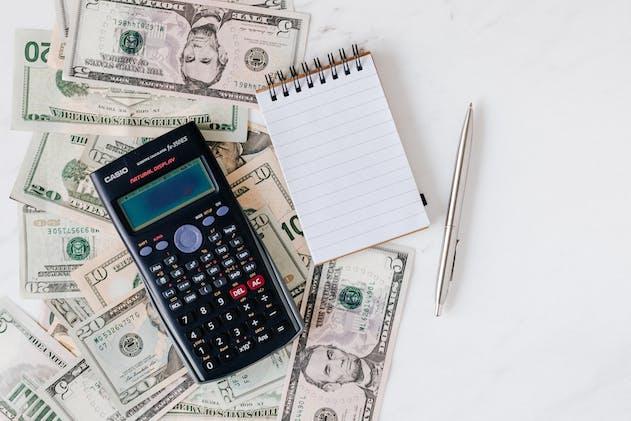 a calculator on top of dollar bills, next to a blank notepad and pen