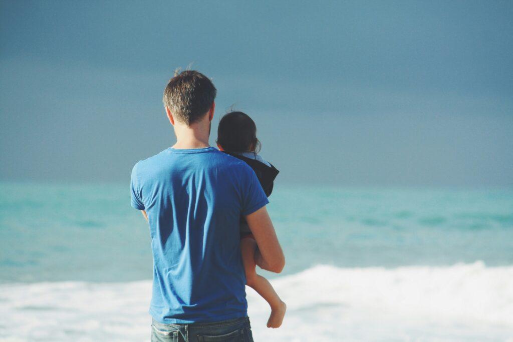 Man standing on a beach holding a small child while looking out at the ocean under an overcast sky.
