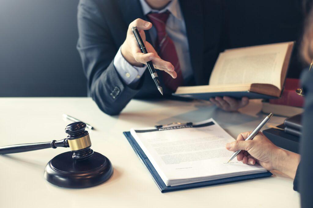 A suited professional gestures with a pen while holding an open legal book, speaking across a desk where another person is signing documents on a clipboard. A judge’s gavel rests nearby, emphasizing the legal and formal setting.