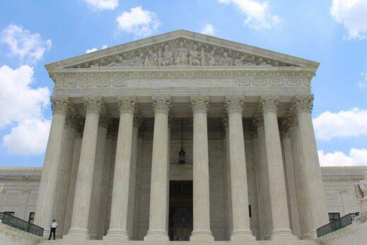 Front view of a grand courthouse with tall marble columns and detailed carvings, set against a bright blue sky with scattered clouds.