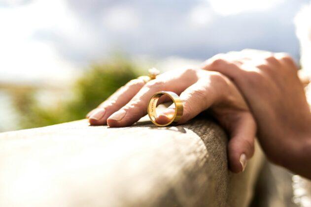 Close-up of a hand sliding off a gold wedding ring on a wooden surface, with soft sunlight and a blurred outdoor background.