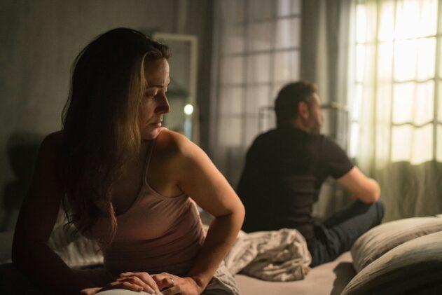 A woman sitting on the edge of a dimly lit bed looking away with a troubled expression, while a man sits turned toward the window in the background, both appearing distant in a tense, emotional moment.