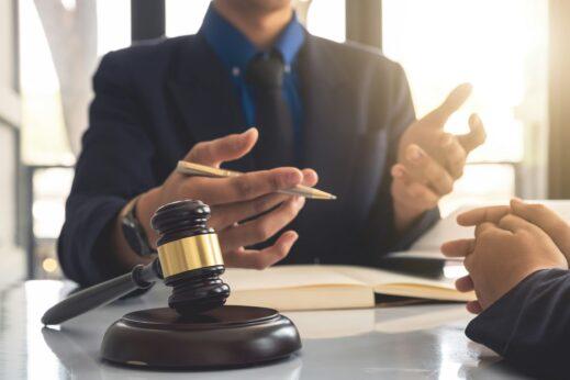 A close-up of a lawyer speaking with a client at a desk, gesturing with a pen while a judge’s gavel and an open notebook sit in the foreground, lit by soft natural light from nearby windows.