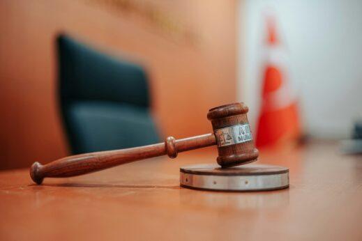 A wooden judge’s gavel resting on its sound block on a polished courtroom table, with a blurred judge’s chair and warm-colored background adding to the formal, legal atmosphere.