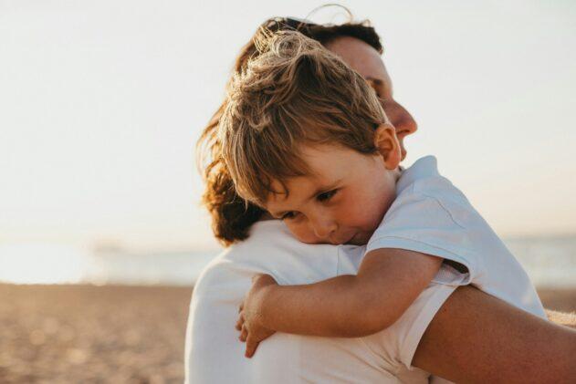 A small child rests their head on an adult’s shoulder while being held close, the two of them standing on a sunlit beach with warm, soft light and the ocean fading into the background.