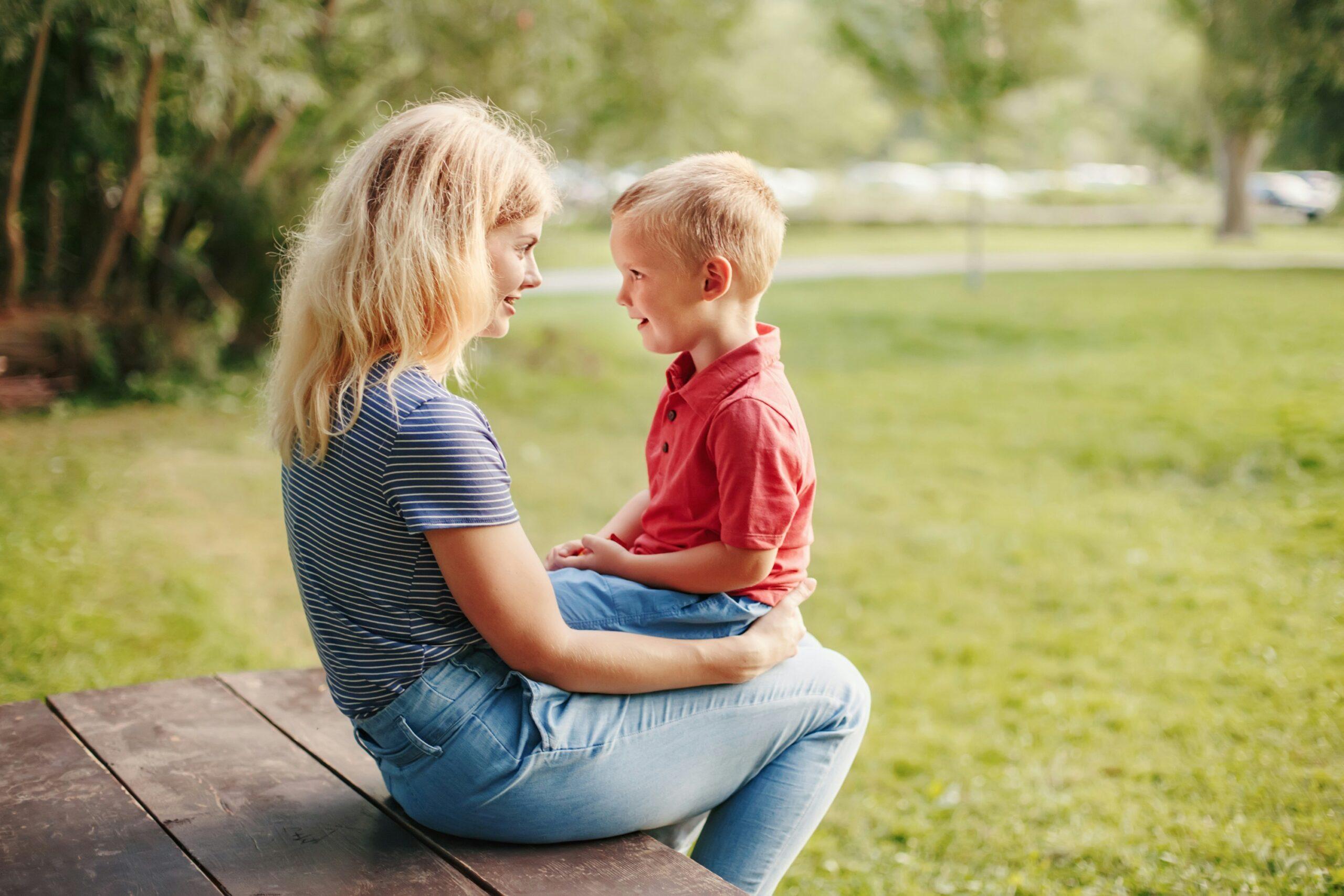 A woman sitting on a wooden bench in a sunny park, gently holding a young boy on her lap as they face each other with warm, engaged expressions, surrounded by soft greenery in the background.