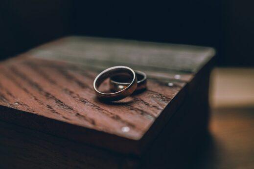 Two metallic wedding bands resting side by side on the edge of a dark wooden surface, photographed in soft, low light with shallow depth of field, emphasizing the rings against a blurred background and creating a quiet, reflective mood.