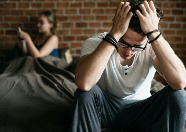 A young man sitting on the edge of a bed with his head in his hands, wearing glasses and a white T-shirt, appearing stressed or overwhelmed, while a woman sits blurred in the background looking at her phone against an exposed brick wall in a bedroom setting.