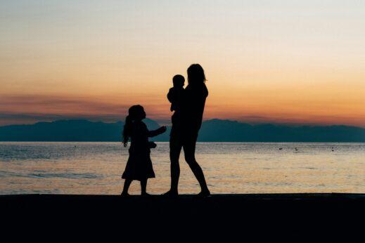 Silhouettes of an adult holding a small child and an older child standing beside them on a beach at sunset, with calm water and distant mountains under a soft orange and blue sky.