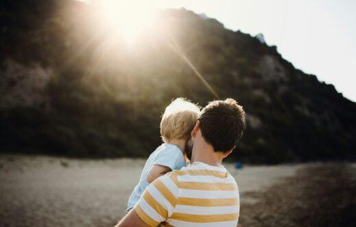 Adult holding a small child while standing on a sandy beach, both seen from behind, facing a sunlit rocky hillside as warm sunlight flares across the scene, creating a calm and intimate outdoor moment.