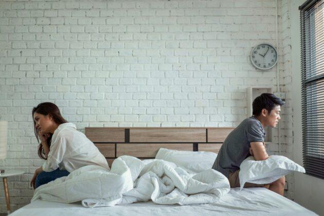 A man and a woman sit back to back on an unmade bed in a bright bedroom, both turned away from each other with tense, withdrawn body language, surrounded by white bedding, a brick wall, a wall clock, and window blinds letting in daylight.
