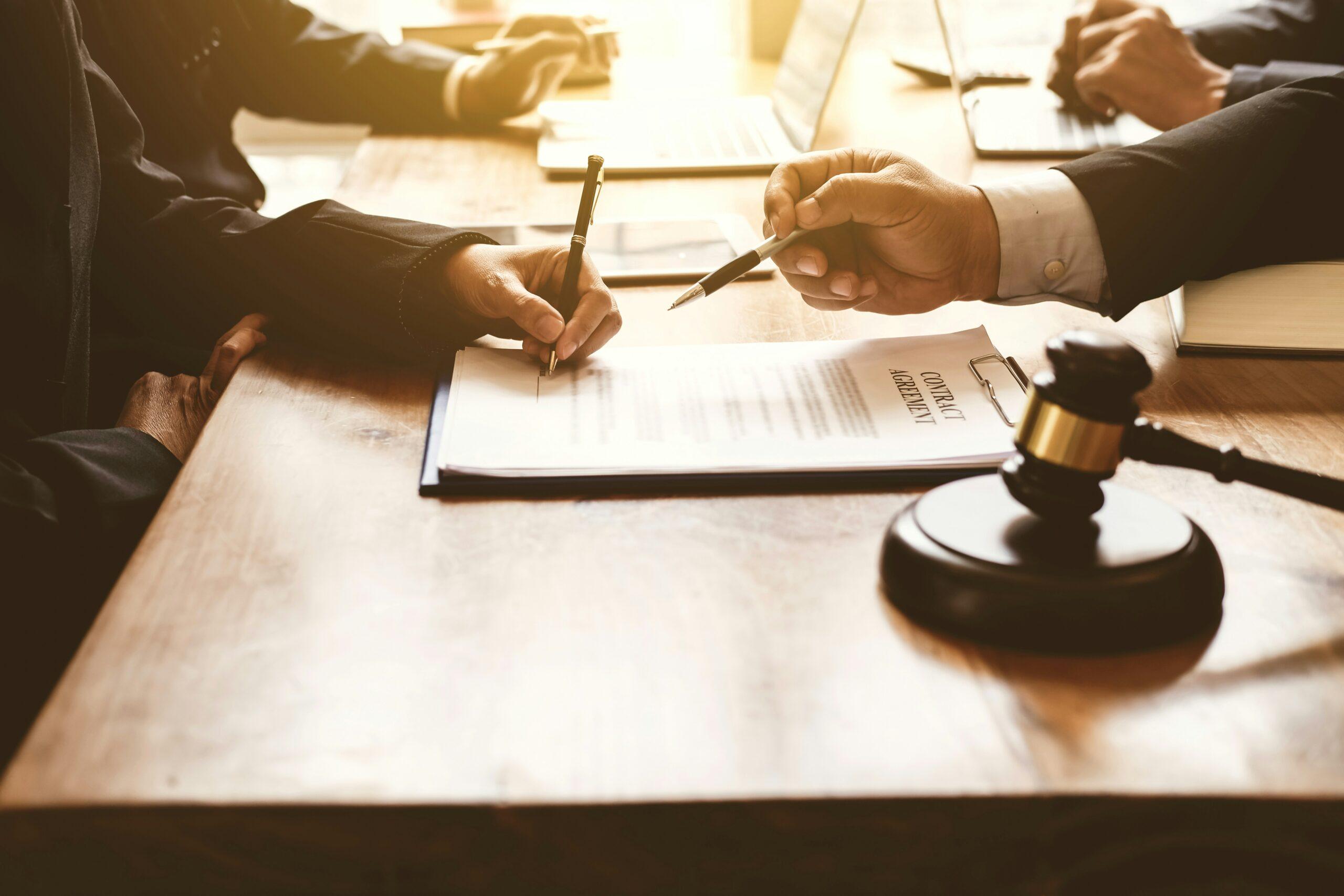 Two people seated at a wooden table review and sign a printed legal contract, each holding a pen above the document, with a judge’s gavel resting in the foreground and laptops and paperwork visible in soft, warm lighting.