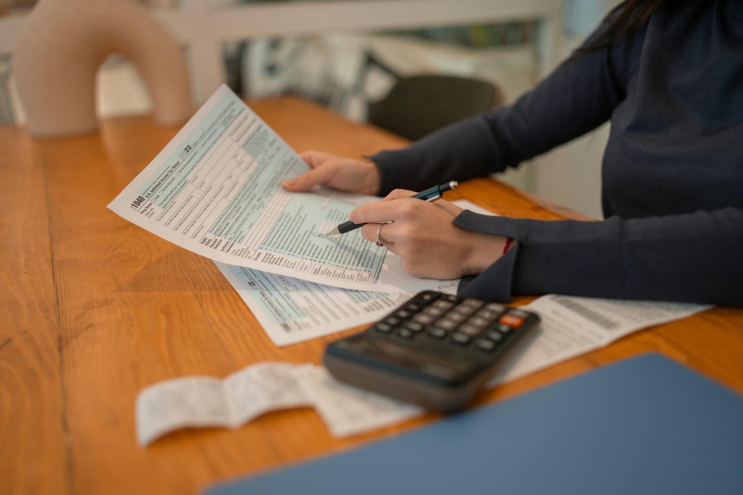 Woman sitting at a desk at home, focused on filling out tax forms with papers and a calculator nearby.