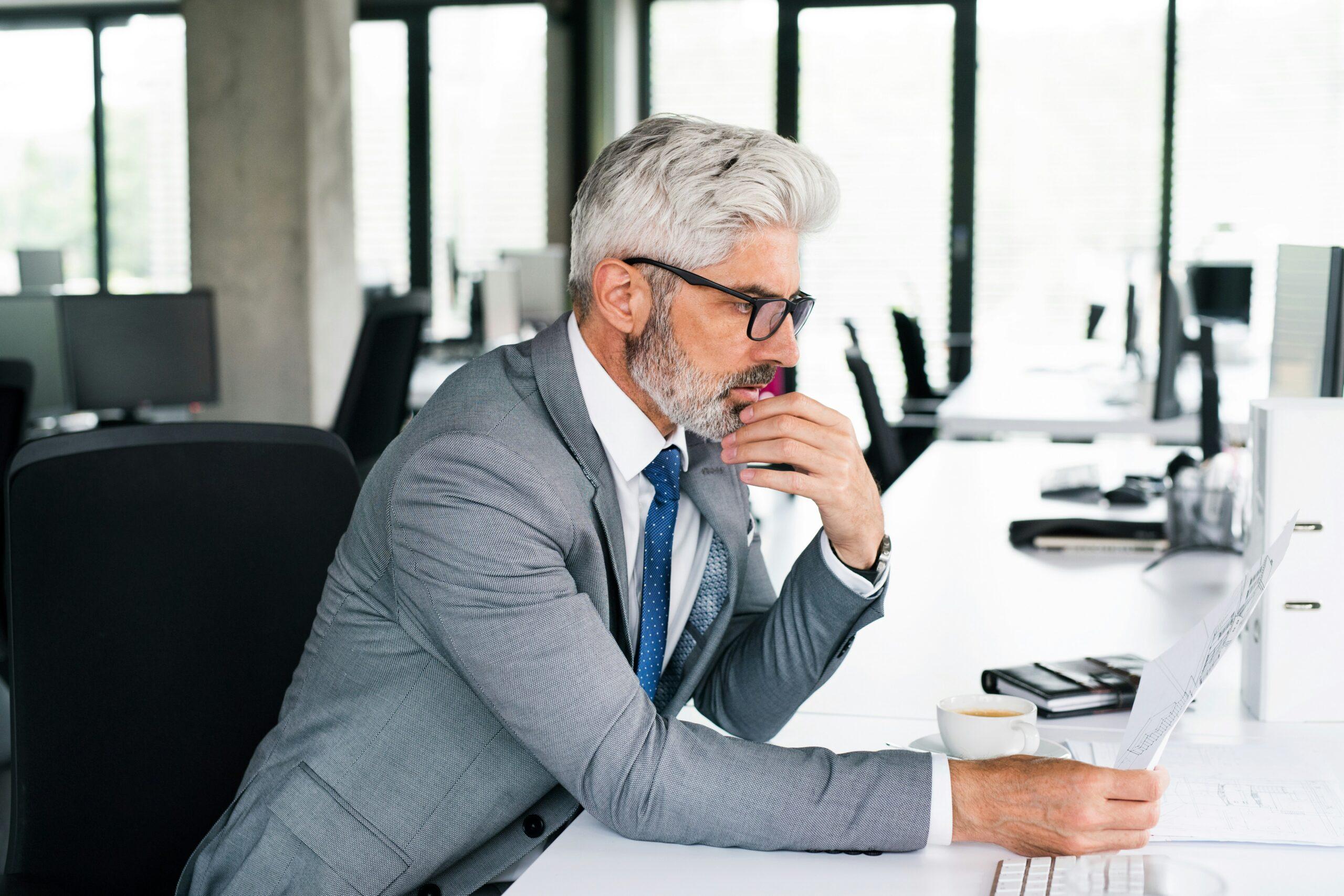 A man in a suit and glasses sits at a desk, working on a laptop.