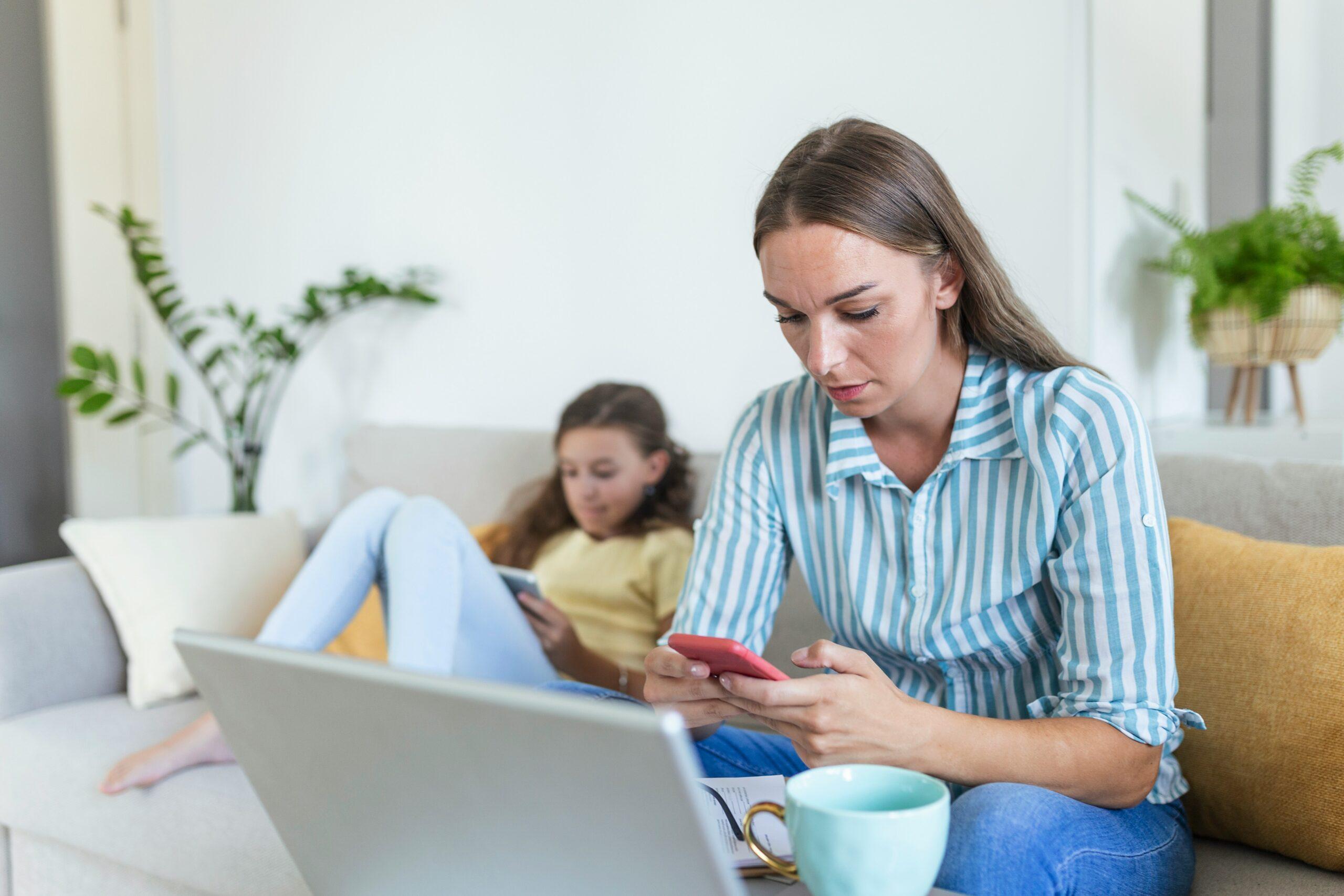 A woman and her daughter sit on a couch, each focused on their cell phones, sharing a quiet moment together.