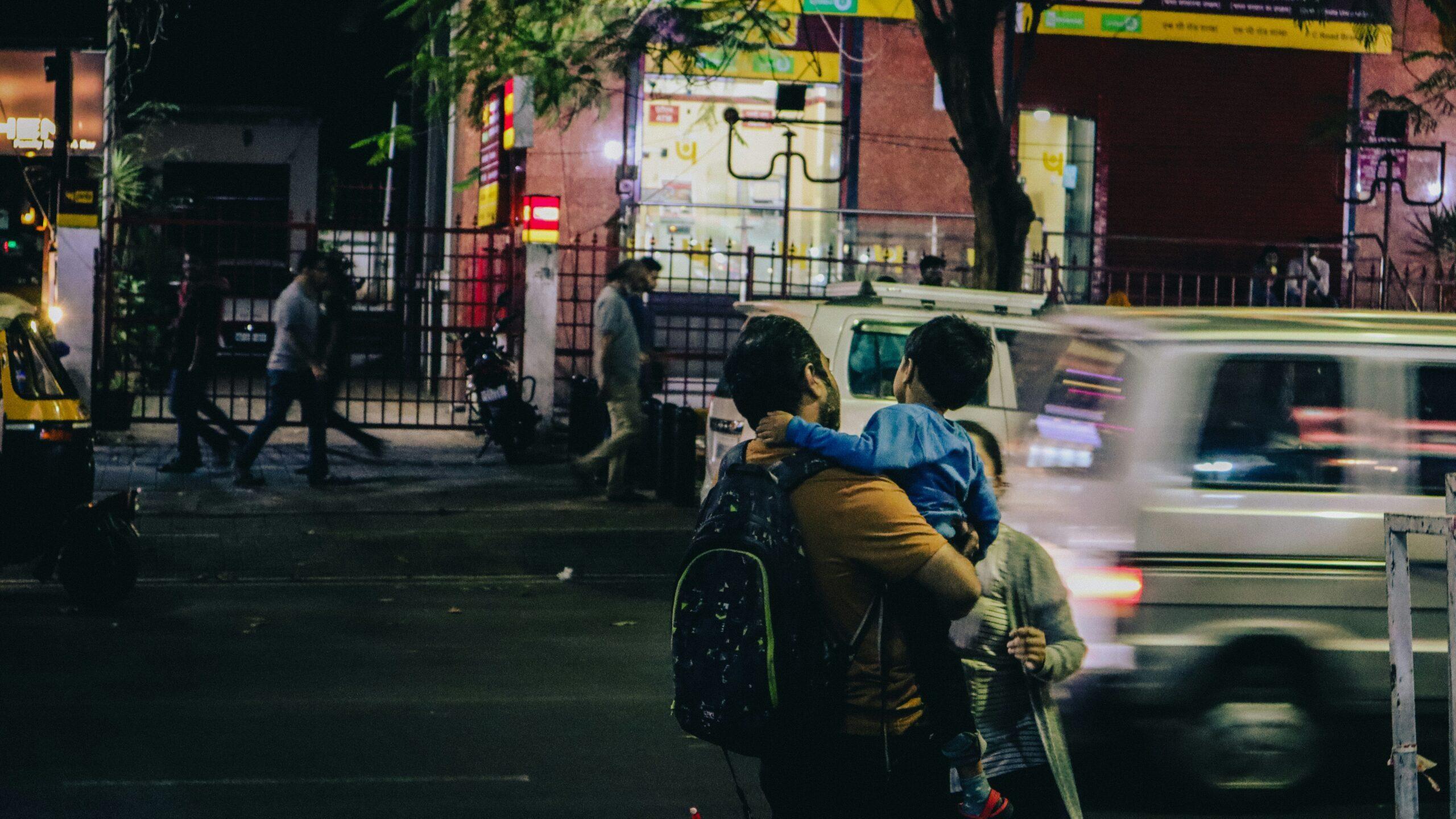 A nighttime urban street scene showing an adult carrying a young child while standing near a busy road, with cars and scooters passing by in motion blur; pedestrians walk along the sidewalk behind a metal fence, illuminated storefront signs glow in the background, and streetlights cast a mix of warm and cool light across the city environment.